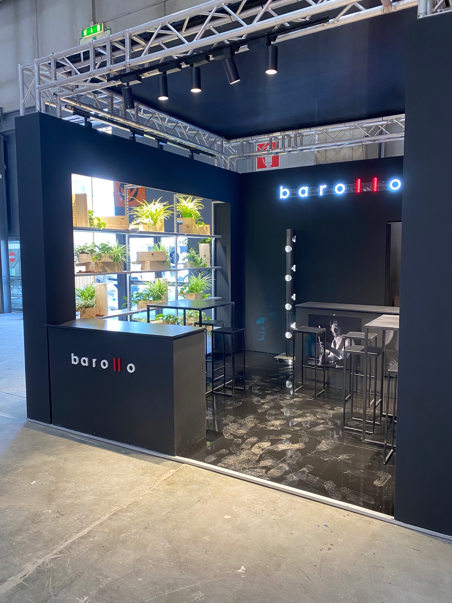 Stand Barollo con banco di accoglienza, tavoli alti, sgabelli e parete espositiva con elementi verdi.
