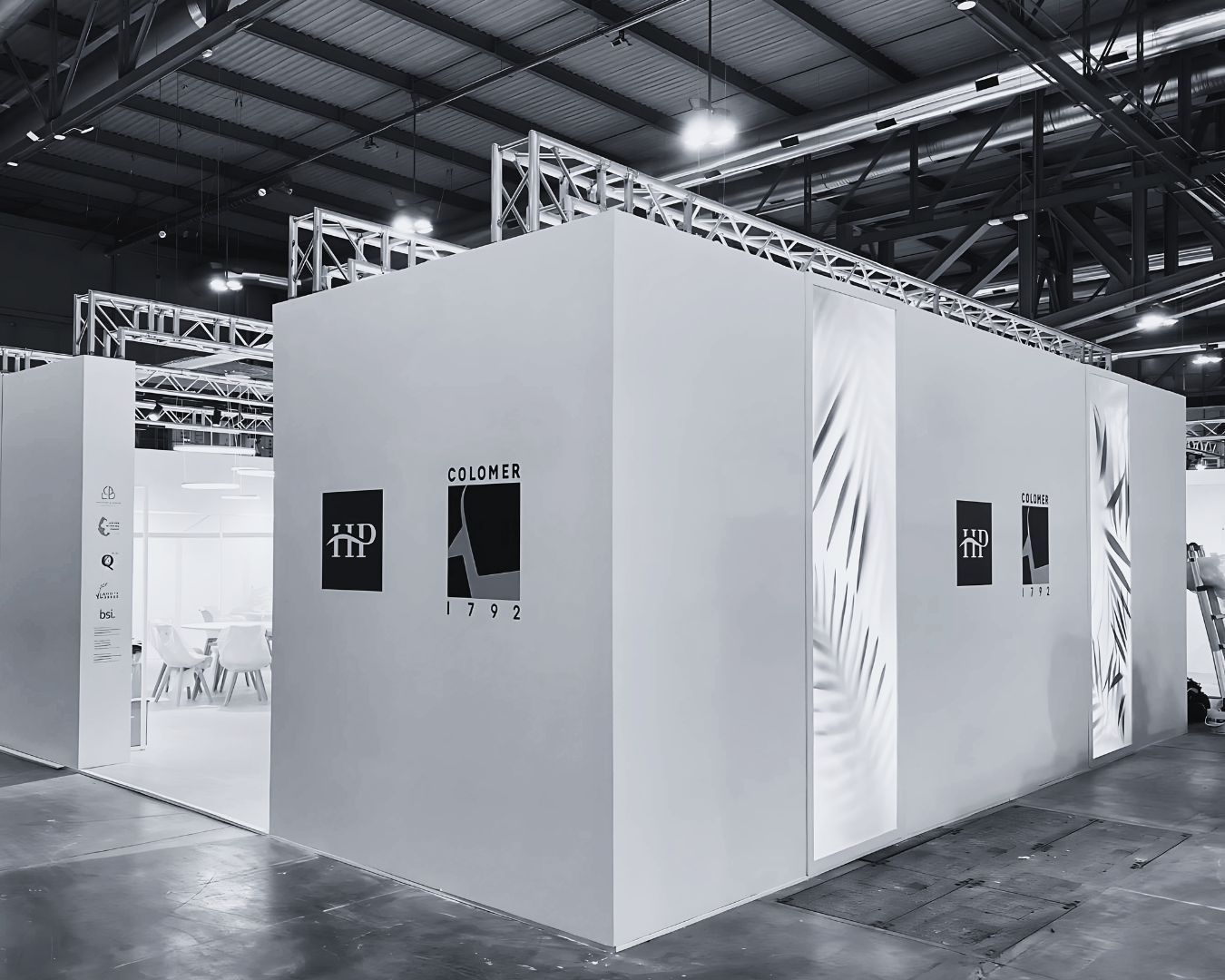 Stand fieristico minimalista con pareti bianche personalizzate e branding Colomer 1792, illuminato da luci d’ambiente.